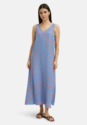 Frau trägt ein ärmelloses blaues Maxikleid mit roten und weißen Blumenmustern und braune flache Sandalen vor weißem Hintergrund.