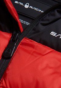 Röd och svart zip-jacka med strukturerad yta, med en framträdande svart dragkedja och "Sail Racing"-logotyp på insidan.