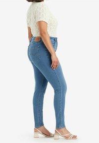 Mujer vestida con jeans ajustados de color azul claro, blusa de punto de manga corta blanca y sandalias de tacón beige, de pie en una pose lateral sobre un fondo blanco.