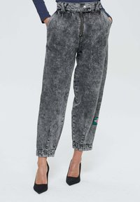 Femme portant un jean gris délavé taille haute et large avec une fermeture éclair frontale, les mains dans les poches, et des escarpins noirs à talons pointus.