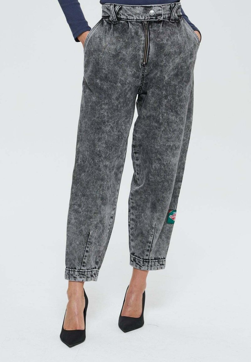 Femme portant un jean gris délavé taille haute et large avec une fermeture éclair frontale, les mains dans les poches, et des escarpins noirs à talons pointus.