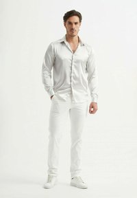 Chemise longue à manches blanches en satin avec un col classique, un devant à boutons et une coupe ajustée, assortie à un pantalon slim blanc et des baskets blanches.