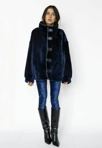 Marineblå imiteret pelsjakke med høj krave og knaplukning, kombineret med skinnende blå leggings og høje, sorte, tætsiddende støvler.