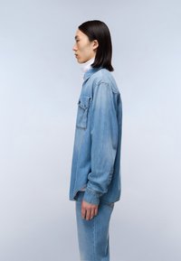 Lyseblå denimskjorte med en enkelt brystlomme, knapstolpe og lange ærmer. Båret over en hvid turtleneck.