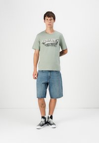 Lys grøn t-shirt med et sort grafisk logo, iført blå denim shorts og sorte højtop sneakers, stående mod en hvid baggrund.