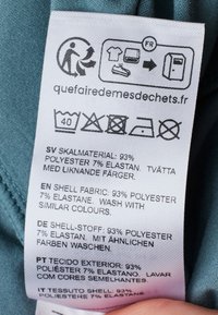 Skötselrådsetikett på ett ljusblått tyg, som visar skötselråd och materialkomposition: 93 % polyester, 7 % elastan, på flera språk.