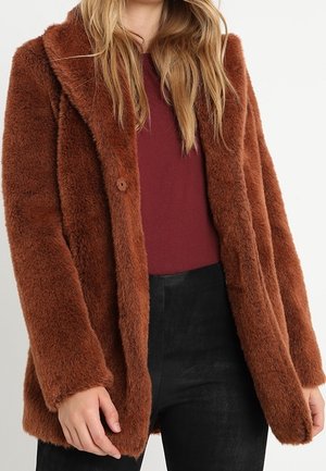 Vrouw draagt een bruine, pluizige jas over een donkerrood topje en zwarte broek, met lang blond haar zichtbaar.