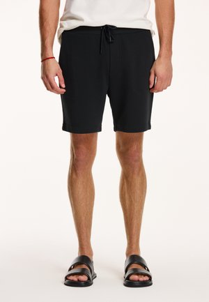 Marineblaue Shorts mit elastischem Bund und Kordelzug, mit entspannter Passform und umgeschlagenen Säumen. Getragen mit schwarzen Rutsch-Sandalen.