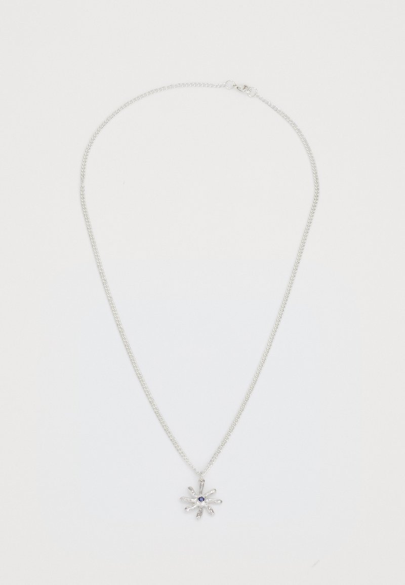 Colar de corrente prata com um pingente em forma de flor, apresentando uma gema azul-escura no centro, sobre um fundo branco liso.