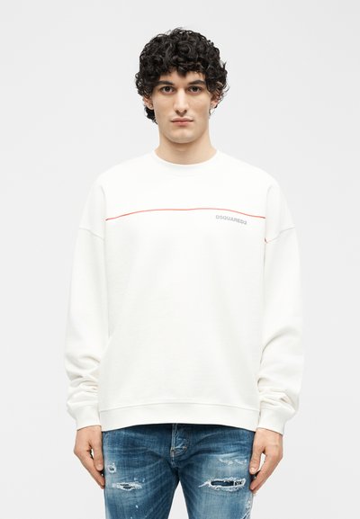 Νέος άνδρας με σγουρά μαύρα μαλλιά, φοράει λευκό φούτερ με κόκκινη επένδυση και ανοιχτό μπλε τζιν με τρύπες, στέκεται μπροστά σε απλό φόντο.