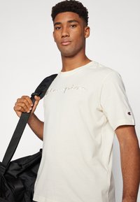 Cremfarbendes T-Shirt mit „Champion“-Logo in gesticktem Text, kurzen Ärmeln und einem rot-weißen Logo-Patch auf dem linken Ärmel.