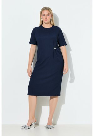 Vrouw die een marineblauwe midi-jurk met korte mouwen draagt en zilveren hoge hakken met grote strikken, staand tegen een witte achtergrond.