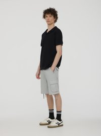 Polo nero abbinato a pantaloni cargo grigi chiari con tasche laterali, orli arricciati, calze a strisce bianche e nere e sneakers beige.