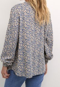 Chemise à manches longues avec un fond beige orné de motifs de feuilles bleues, tissu doux et poignets élastiqués, associée à un jean en denim.