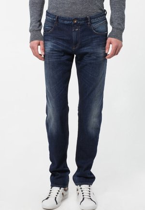 Dark blue denim jeans with a slim fit, five pockets, and subtle fading near the thighs, paired with white sneakers.