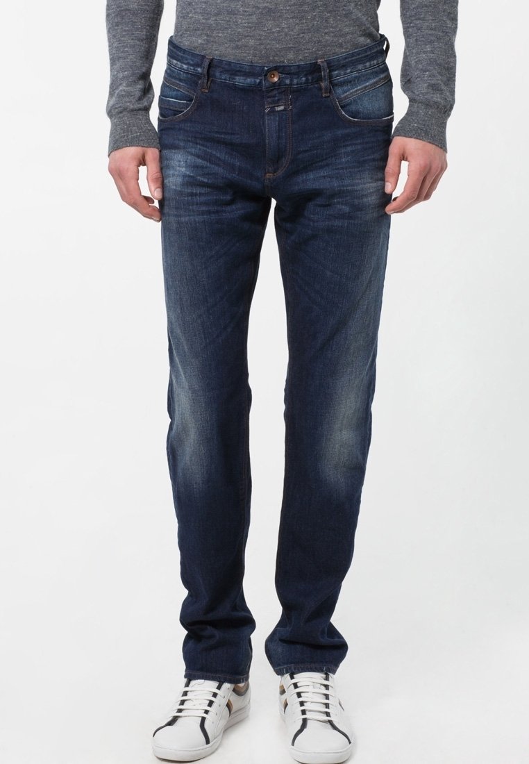 Mörkblå denimjeans med slim fit, fem fickor och subtila blekningar vid låren, tillsammans med vita sneakers.