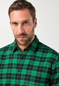Camisa de franela a cuadros verdes y negros con cuello abotonado y botones negros, que presenta una textura de tela suave y un patrón clásico.