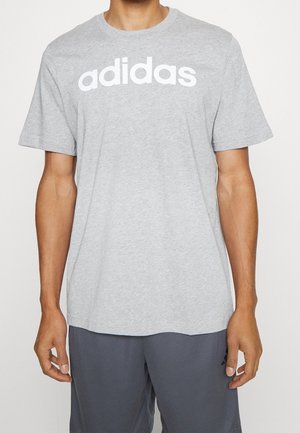 Mies pukeutuneena vaaleanharmaaseen Adidas-paitaan, jossa on valkoinen logoteksti, ja tummanharmaisiin shortseihin, seisoo yksinkertaisen valkoisen taustan edessä.