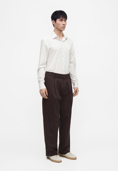 Jeune homme debout portant une chemise blanche boutonnée, un pantalon brun foncé et des chaussures beige clair sur un fond blanc uni.