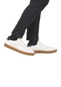 Sneaker in pelle bianca con accenti neri sul tallone, caratterizzate da una superficie liscia e una suola in gomma, presentate con pantaloni in denim scuro.