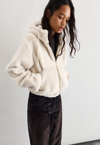 Kremna umetna krzna jakna z zadrgo in kapuco, ki ima tudi vrvico, stranske žepe in krajšo zasnovo, v paru z temnimi kord pantami.