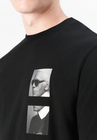 Fekete pamut póló, amelyen egy napszemüveges figura kettős grafikai nyomat található, kerek nyakkal és rövid ujjakkal.