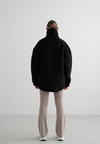 Personne debout, dos tourné vers l'avant, portant une veste polaire noire oversize, un pantalon beige et des baskets blanches.