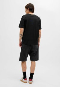 Sort bomulds t-shirt med en afslappet pasform, båret med mørke denim shorts. Viser synlige tatoveringer og afslappet fodtøj med stropper og røde accenter.