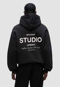 Černá mikina s kapucí ve volném střihu, se velkým bílým textem na zadní straně, který zní "WRSTBHVR STUDIO GARMENTS" a dalšími detaily níže.