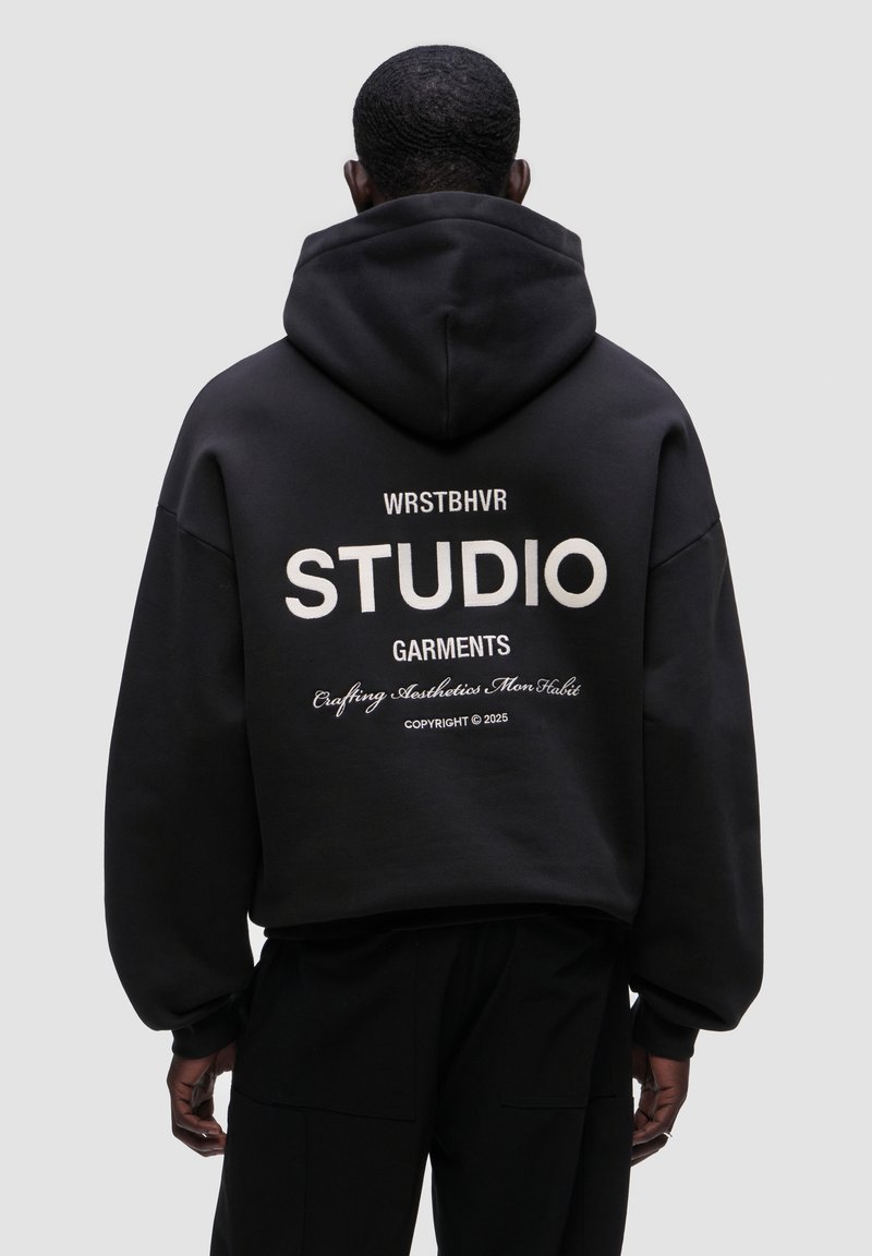 Černá mikina s kapucí ve volném střihu, se velkým bílým textem na zadní straně, který zní "WRSTBHVR STUDIO GARMENTS" a dalšími detaily níže.