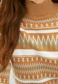 Brązowy sweter w wzory z prążkowanym, błyszczącym kołnierzem typu golf. Ma geometryczne wzory w kolorze białym, różowym i zielonym na całej powierzchni.