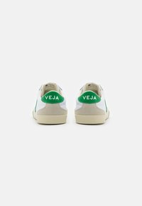 Baltie sneakers ar zaļiem akcentiem, ar gludu virsu, zamšādas papēža cilpiņu un teksturētu gumijas zoli. Logo redzams uz papēža.
