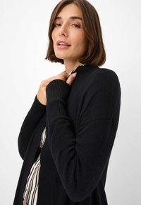 Schwarze Strickjacke mit gerippten Bündchen, offenem Vorderteil und einer weichen Textur. Das Model hält das Kleidungsstück nah, trägt ein gestreiftes Oberteil darunter.