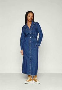 Mujer con trenzas que lleva un vestido largo de mezclilla abotonado, con un lazo en la cintura y zapatillas altas amarillas mostaza, de pie contra un fondo liso.