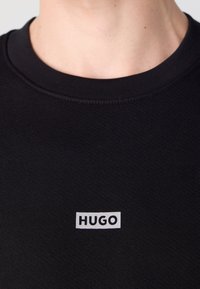 Primer plano de una camiseta negra de cuello redondo con un logo rectangular blanco que dice "HUGO" centrado en el pecho.