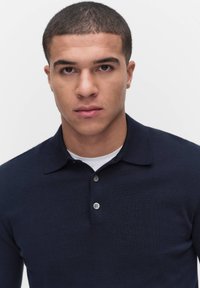 Tummansininen pitkähihainen polo-paita, jossa on ribbipinta, tasainen kaulus ja kaksi nappia, päällä valkoinen pyöreäkauluksinen paita.