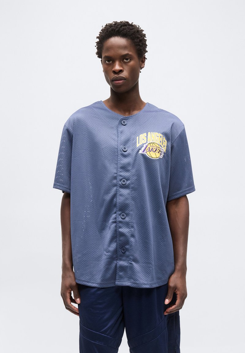 Fiatal férfi, aki kék Los Angeles Lakers hálós gombos mezben és hozzá illő sötétkék nadrágban áll, egyszerű háttér előtt.