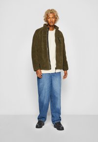 Giacca in fleece verde oliva con colletto alto, zip frontale e tasca nera a contrasto. Abbinata a jeans blu larghi e sneakers nere.
