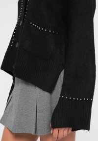 Cardigan noir en tissu texturé, avec des accents en studs métalliques le long des poches et des manches. Porté sur une jupe à carreaux grise.
