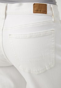 Jeans de mezclilla blanca con una etiqueta de cuero etiquetada "American Eagle", bolsillos traseros y detalles de costura sutiles. Textura suave y diseño ajustado.