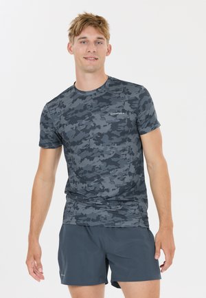 Junger Mann, der ein graues Tarn-Trainingsshirt und dunkelgraue Shorts trägt, vor einem schlichten weißen Hintergrund stehend.