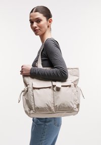 Sac fourre-tout beige en tissu léger avec deux poches avant, détails de cordon de serrage réglables et bretelles robustes.