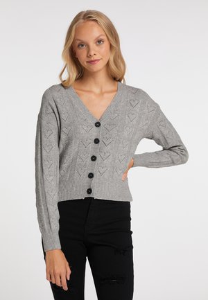 Blonde Frau trägt eine graue V-Ausschnitt-Strickjacke mit Herzmustern und schwarze zerrissene Skinny-Jeans und posiert mit einer Hand auf der Hüfte.