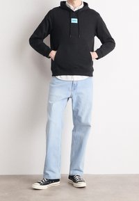 Sudadera negra con capucha y un pequeño logo azul, bolsillo delantero, combinada con vaqueros de pierna recta azul claro y zapatillas de lona negras.