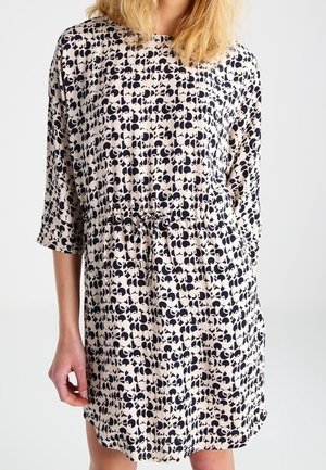 Femme portant une robe beige avec un motif abstrait noir, manches trois-quarts et taille à cordon, debout devant un fond uni.