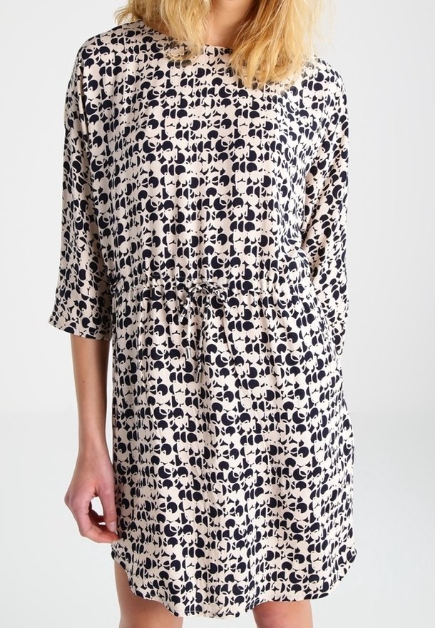 Femme portant une robe beige avec un motif abstrait noir, manches trois-quarts et taille à cordon, debout devant un fond uni.