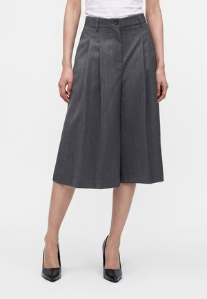 Kobieta nosząca szerokie, szare culottes z fałdami, połączone z czarnymi szpilkami z ostrym noskiem, stojąca na tle jednolitego białego tła.