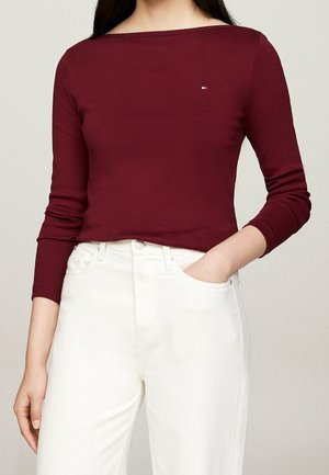 Mujer con una camiseta ajustada de manga larga color burdeos con un pequeño logo y pantalones blancos de talle alto, con la mano en el bolsillo.