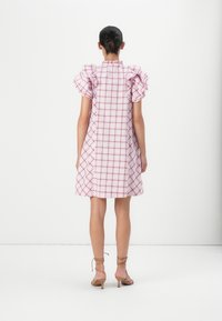 Femme avec les cheveux attachés portant une robe à carreaux rose et blanche jusqu’aux genoux avec des manches courtes bouffantes et des sandales à talons couleur beige, debout, dos visible.