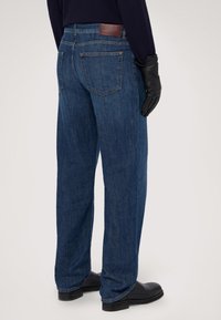 Vaqueros de mezclilla azul con corte recto, que cuentan con bolsillos traseros y una etiqueta de cuero marrón en la cintura. Llevados con guantes y botas negras.